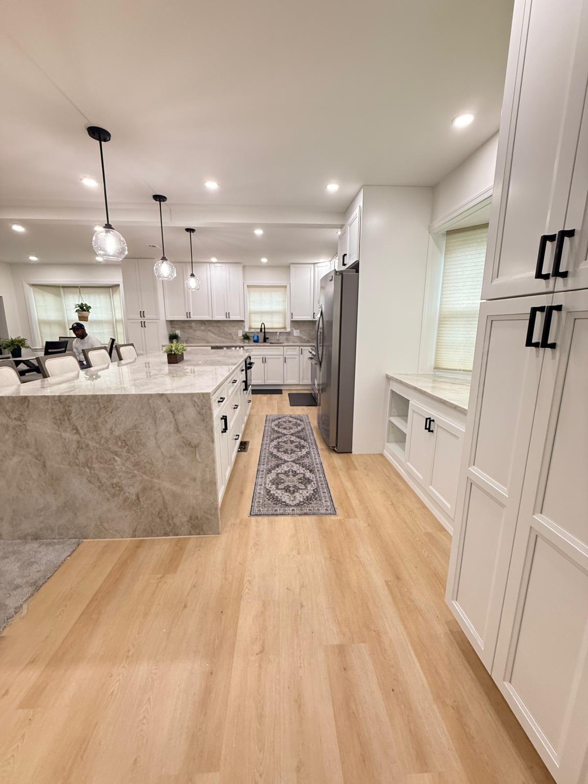 Finished kitchen seen down the aisle between a waterfall-edge island and tall pantry cabinets with light wood-look flooring