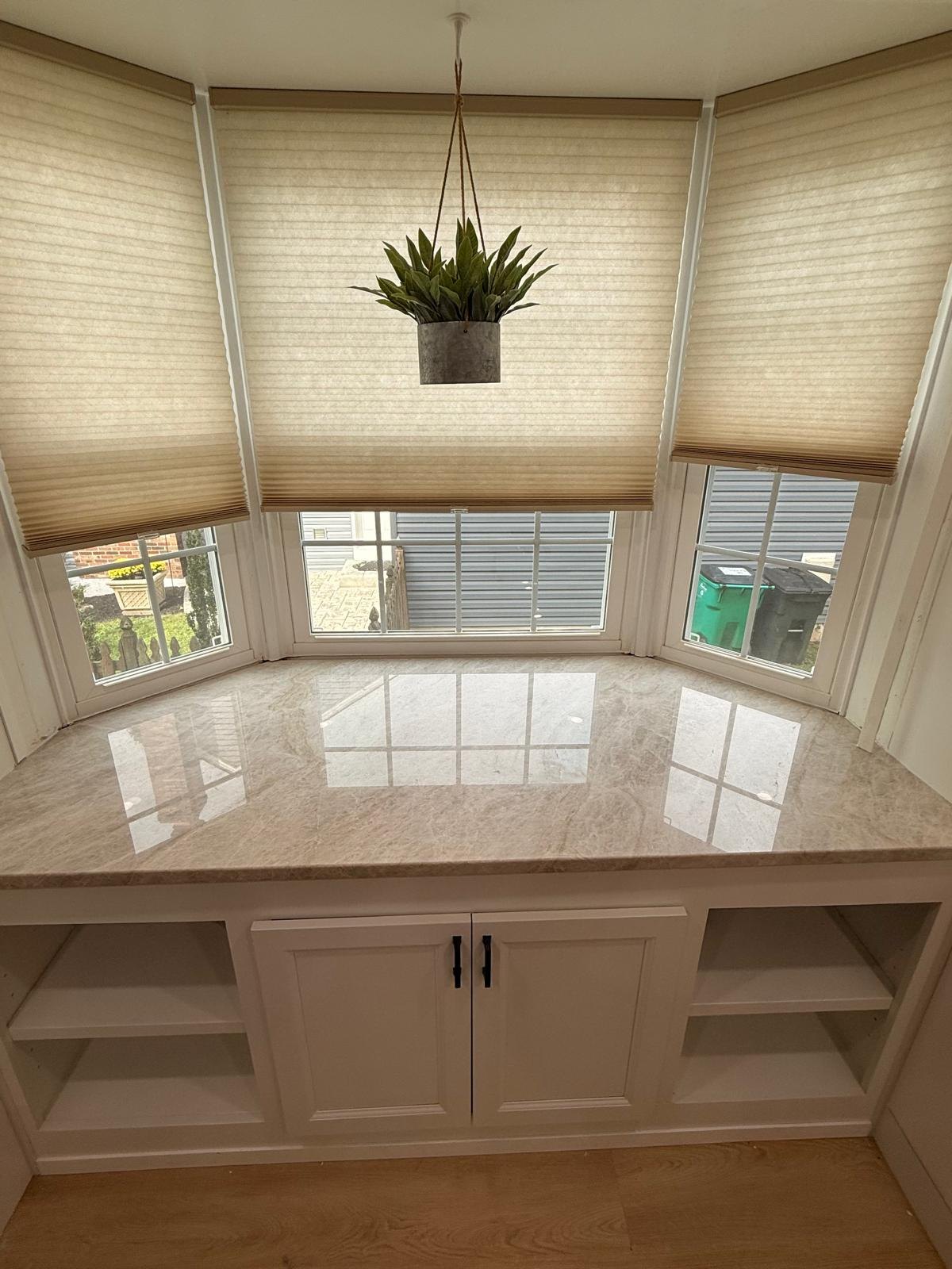 Finished kitchen bay-window bench with white shaker cabinets, open side shelves and a polished stone countertop under a pleated shade