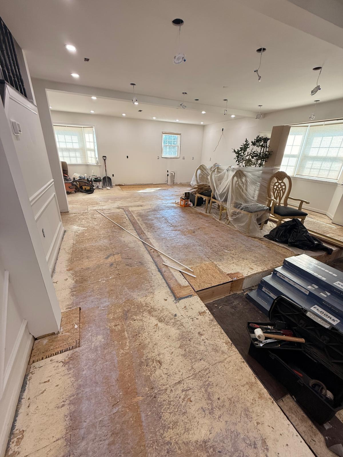 Open living and dining area with patched subfloor, tools on site and stacked click-lock flooring boxes
