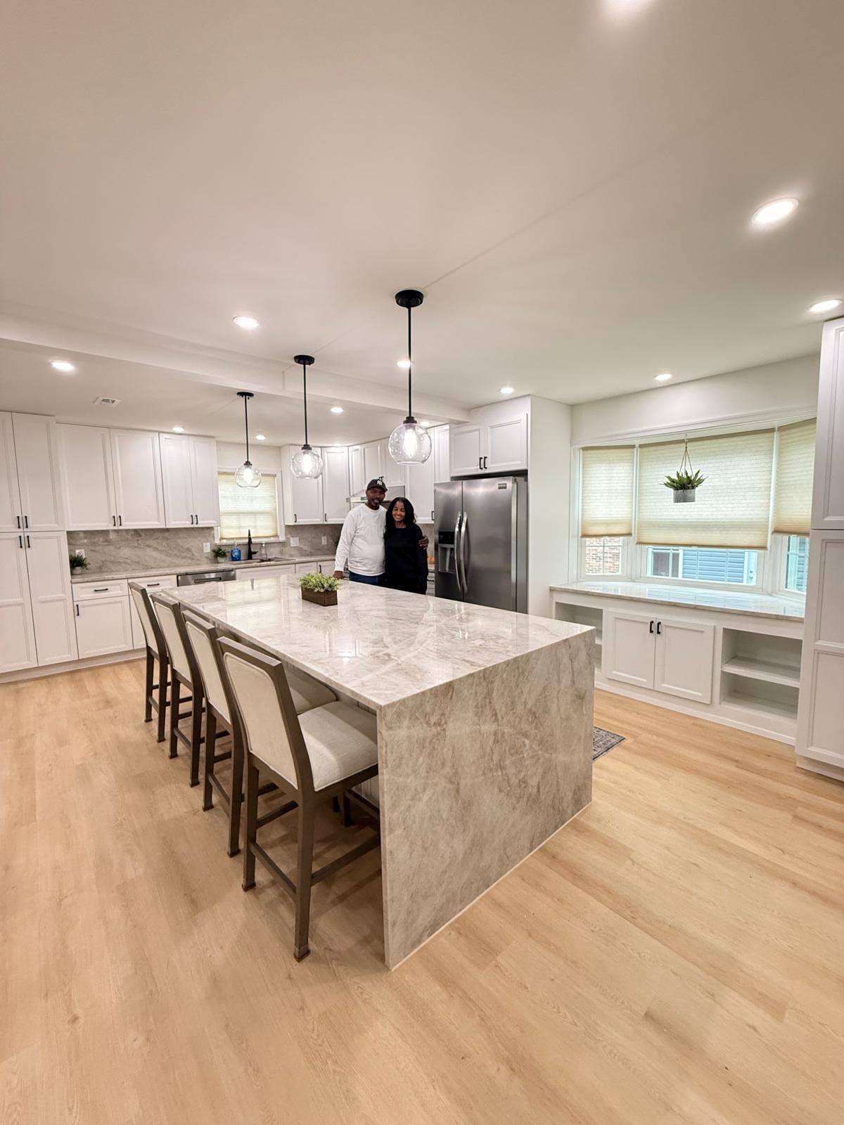 Two smiling homeowners standing behind the finished kitchen island with a waterfall stone countertop and glass globe pendants