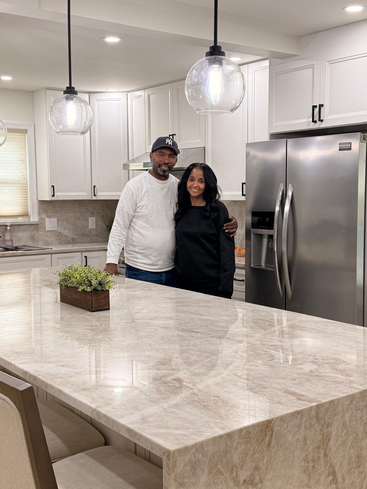 Close-up of two smiling homeowners at their finished kitchen island with white cabinets, a stainless refrigerator and glass pendant lights