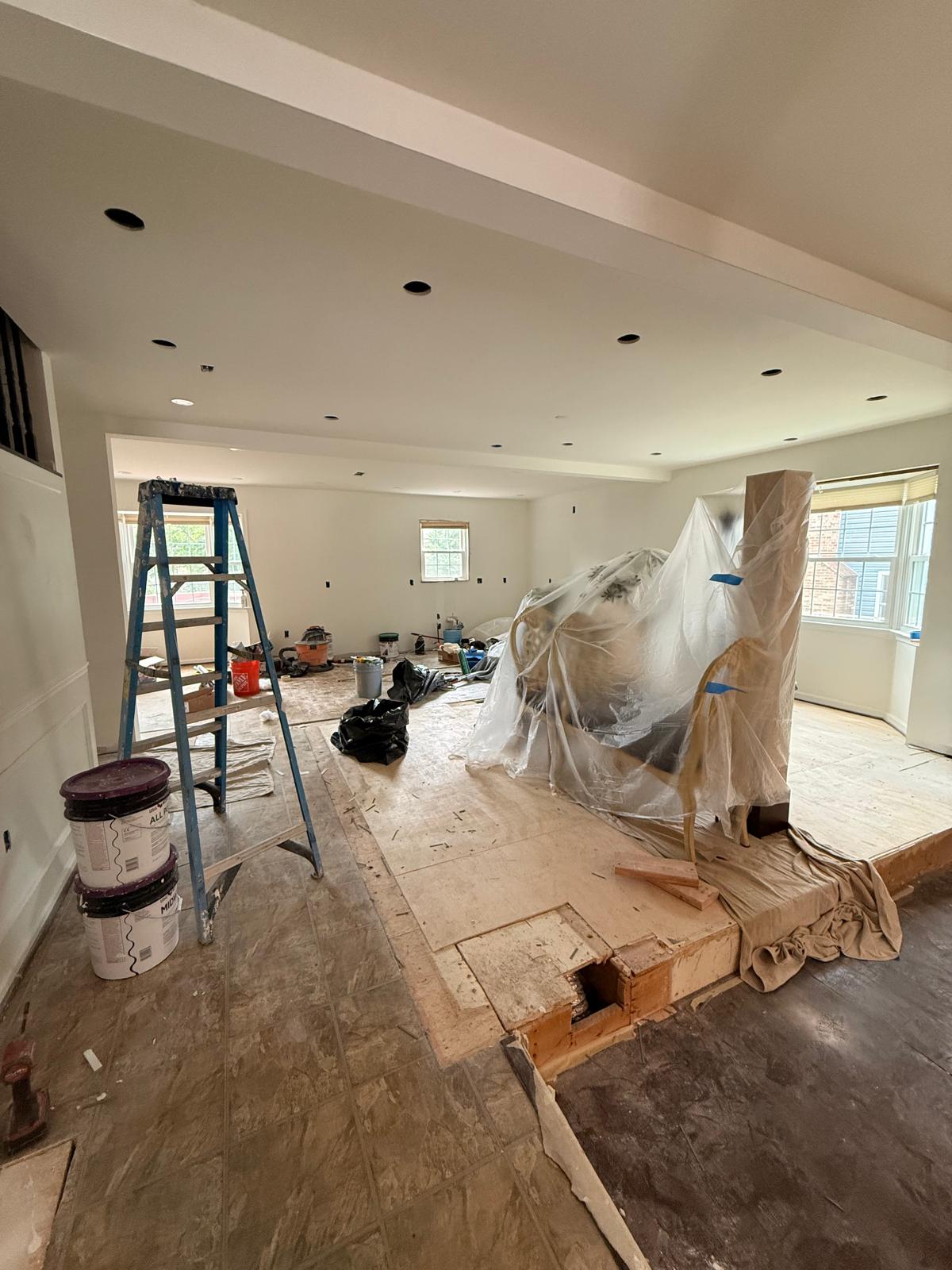 Open-concept living and dining room with old flooring removed down to the plywood subfloor and furniture wrapped in protective plastic